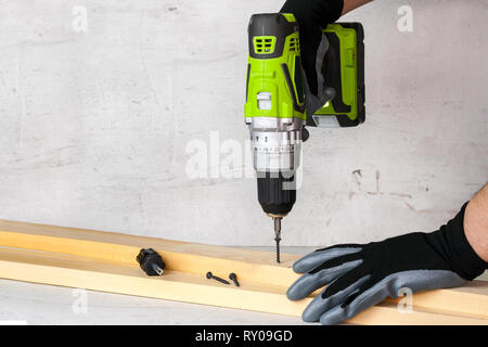 Le constructeur tient dans sa main un tournevis électrique sur l'arrière-plan d'un mur de béton. Vis vis dans une poutre en bois. DIY. Banque D'Images