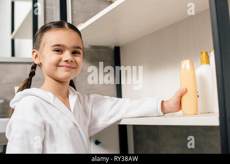 Cute child en peignoir blanc gel douche prise à partir de la tablette dans salle de bains Banque D'Images