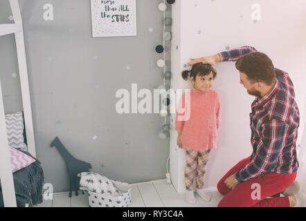 Son père est la mesure de la hauteur de bébé sur le mur. Jolie petite fille et son jeune et beau père jouent ensemble dans la chambre de l'enfant Banque D'Images