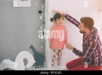 Son père est la mesure de la hauteur de bébé sur le mur. Jolie petite fille et son jeune et beau père jouent ensemble dans la chambre de l'enfant Banque D'Images