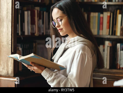 Étudiant intelligent girl, debout près de la fenêtre à la bibliothèque et de la lecture pour l'examen du livre Banque D'Images