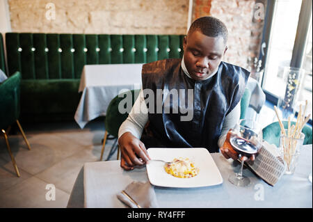 African man in black vêtements traditionnels et de manger des pâtes et boire du vin. Banque D'Images