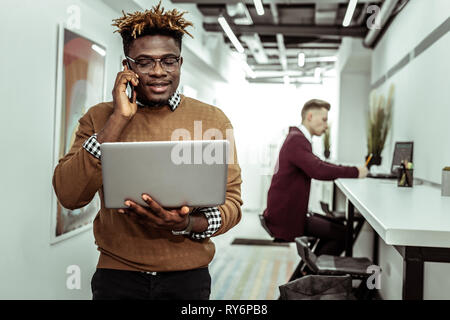 La peau foncée concentré guy répondre au téléphone tout en exerçant son ordinateur portable Banque D'Images