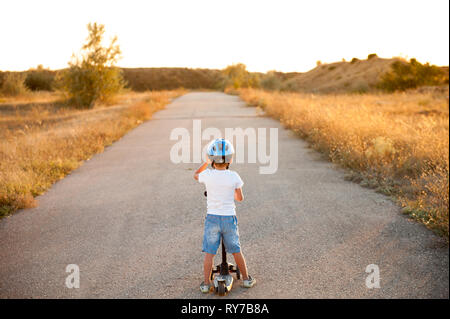 Petit garçon sain et actif en casque et denim shorts debout sur la route vide avec scooter dans le coucher du soleil Banque D'Images
