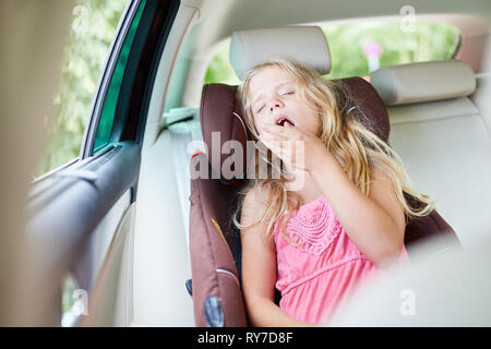 Fille dans la voiture dans le siège enfant sur le siège arrière bâille fatigué sur le voyage Banque D'Images