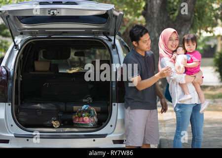 Jeune famille musulmane , transports, loisirs, road trip et personnes concept - heureux homme, femme et petite fille jouant à la poupée à côté de voiture avec trunk ope Banque D'Images