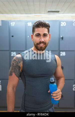 Portrait of smiling homme confiant holding water bottle debout contre les casiers en salle de sport Banque D'Images