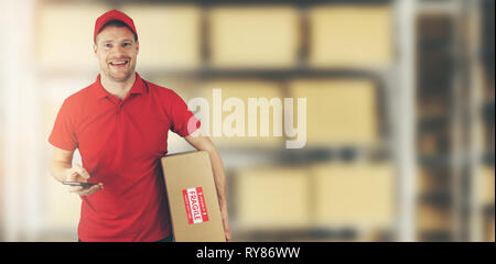 Delivery man standing in warehouse holding cardboard box et téléphone mobile Banque D'Images