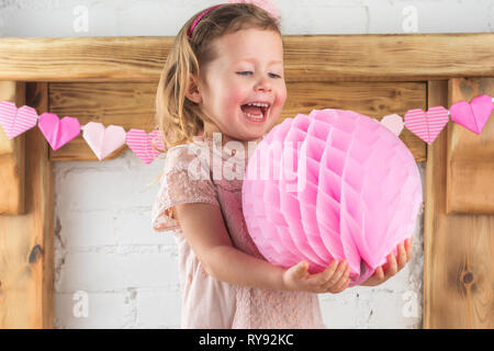 Petite fille en robe rose pastel et bandeau d'abeilles holding pom pom rire avec bonheur, le bois massif de la cheminée background, selective focus Banque D'Images