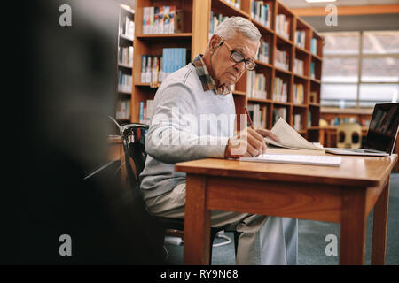 Man studying in classroom. Vieil homme écrit dans un livre assis en classe avec un ordinateur portable à l'avant. Banque D'Images