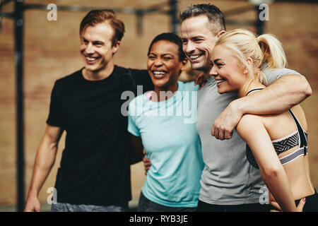 Groupe diversifié de laughing friends in sportswear standing arm in arm dans une salle de sport après une séance de classe ensemble Banque D'Images
