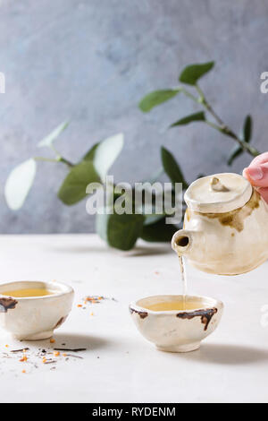 La coulée de verre de thé vert chaud dans deux chinois traditionnels tasse en céramique argile debout sur la table en marbre blanc. Avec branche verte à l'arrière-plan. Banque D'Images