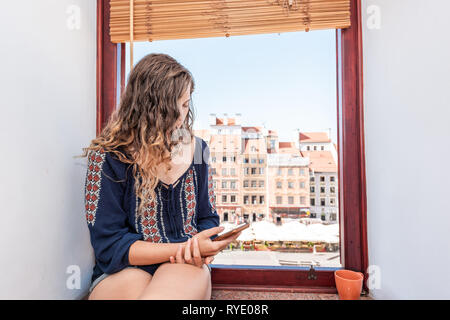 Young woman looking at phone assis sur un rebord de fenêtre de l'appartement avec vue sur la place du vieux marché de la ville de Varsovie, Pologne Banque D'Images