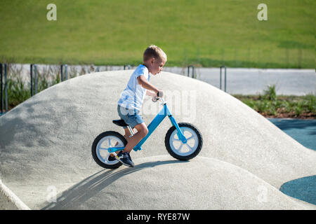 Un petit garçon jouit d'un équilibre vélo sur collines artificielles sur une aire de jeux dans un parc sur une journée ensoleillée d'été. Banque D'Images