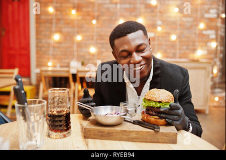 Young african american man respectable en costume noir et des gants pour l'alimentation sitting in restaurant avec double savoureux burger et boire de soude. Banque D'Images
