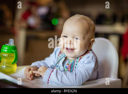 Cute baby girl sitting in chaise haute, manger du pain and smiling Banque D'Images
