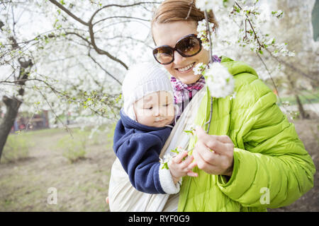 Jeune femme portant son bébé fille dans le tissu pour envelopper l'extérieur dans le parc au printemps. Maman et bébé vers de blooming twig. Banque D'Images