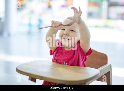 Jolie petite fille assise dans une chaise haute, en tendant les mains avec fourchette et rire Banque D'Images