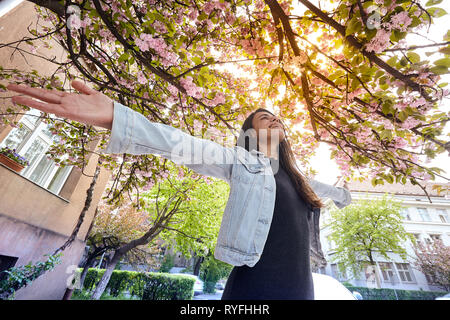 Jeune fille avec les bras grand ouverts et profiter du soleil en plein air permanent dans la lumière du soleil sur l'arrière-plan de sakura en fleurs Banque D'Images