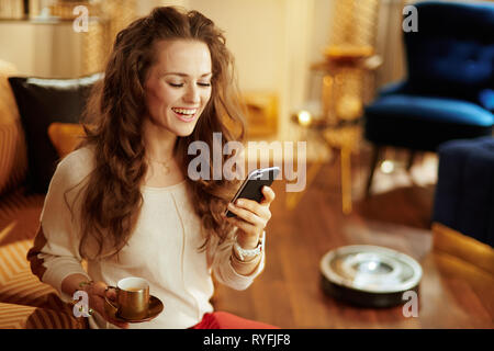 Happy housewife avec de longs cheveux brune avec une tasse à café à l'aide de smart home app et robot-aspirateur nettoyage planchers en arrière-plan dans la maison moderne. Banque D'Images