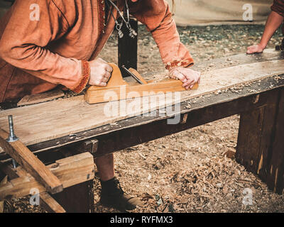 Carpenter à l'époque médiévale vêtements coton Travailler avec un bois en avion. L'homme s'accroche manuellement une planche en bois avec un avion. Construction d'une écorce de bois Banque D'Images