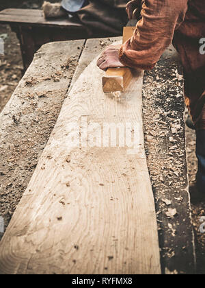 Carpenter à l'époque médiévale vêtements coton Travailler avec un bois en avion. L'homme s'accroche manuellement une planche en bois avec un avion. Construction d'une écorce de bois Banque D'Images