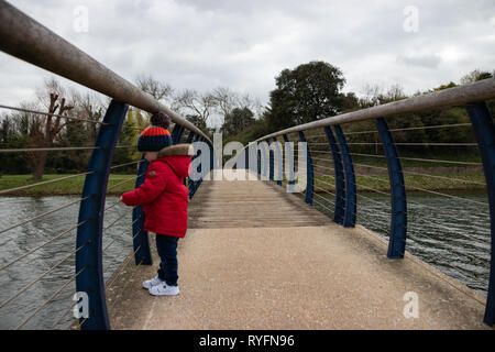 Deux ans en veste rouge et bobble hat sur un pont en bois à la recherche sur le côté au lac ci-dessous Banque D'Images