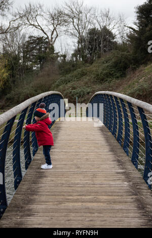 Deux ans en veste rouge et bobble hat sur un pont en bois à la recherche sur le côté au lac ci-dessous Banque D'Images