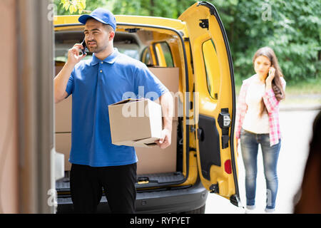 Homme portant un uniforme bleu livraison Holding boîte en carton et parlant au téléphone. Van ouvert en arrière-plan. Banque D'Images