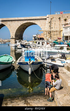 Trois garçons jouer près de la pêche bateaux dans le petit port du Vallon des Auffes à Marseille, France, avec le pont-route dans l'arrière-plan. Banque D'Images