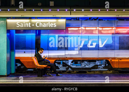 Un homme en costume noir regarde son smartphone dans la gare Saint-Charles à Marseille, France, assis en face d'une peinture murale d'un TGV. Banque D'Images