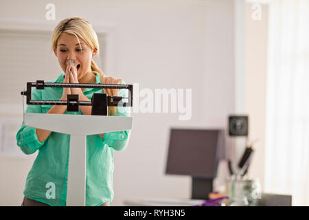 Les jeunes excités femme pesant elle-même sur une échelle de poids médical dans un cabinet de médecin. Banque D'Images