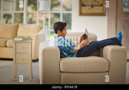 Jeune garçon assis avec ses pieds jusqu'à un fauteuil à l'aide d'une tablette numérique. Banque D'Images