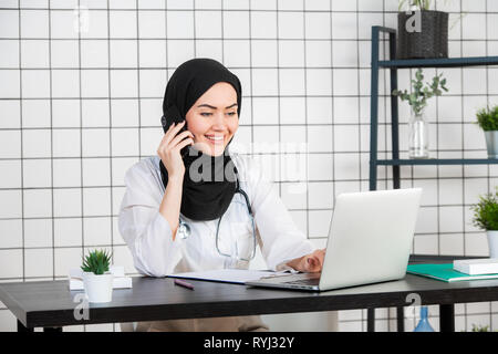 Femme scientifique voilée assise sur son bureau avec les doigts sur un clavier de son ordinateur portable, en souriant. Banque D'Images