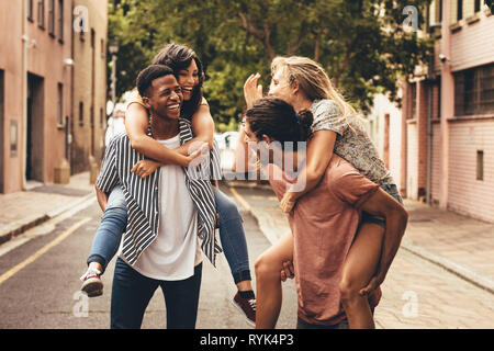 Deux jeunes hommes donnant leurs copines piggyback ride. Cheerful young woman outdoors on city street. Banque D'Images