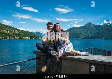 Heureux couple siège avec deux verres d'Aperol spritz, avec vue panoramique sur le lac, les collines et montagnes enneigées à Côme, en Italie. Smiling guy avec ma copine. Banque D'Images