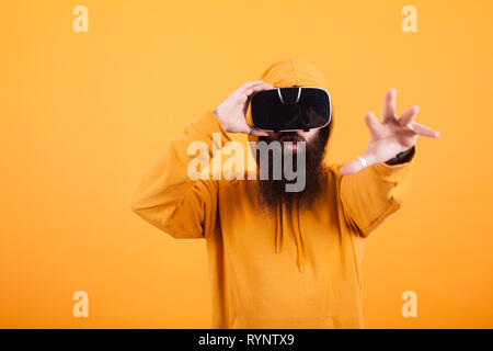Homme barbu portant casque de réalité virtuelle s'adressant avec sa main sur fond jaune. Sweat jaune. Bel homme. Homme hipster. Banque D'Images