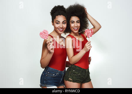 Sweet beauty. Deux belles jeunes afro-américain les femmes dans des vêtements d'été tenue en forme de sucettes coeur et sourire tout en se tenant à l'arrière-plan gris Banque D'Images