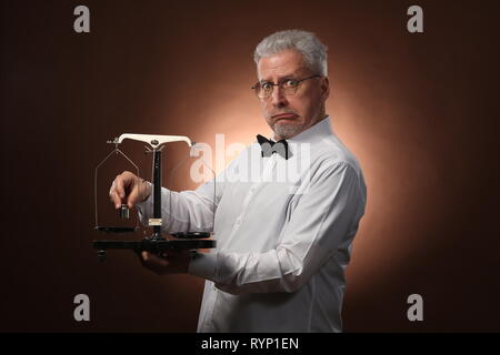 Homme aux cheveux gris âgés de 50 ans, en chemise blanche, lunettes et chapeau pesant quelque chose sur les balances avec petit kettlebells Banque D'Images