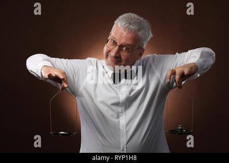 Homme aux cheveux gris âgés de 50 ans, en chemise blanche, lunettes et chapeau pesant quelque chose sur les balances avec petit kettlebells Banque D'Images