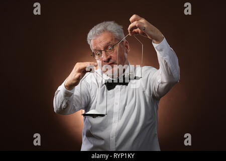 Homme aux cheveux gris âgés de 50 ans, en chemise blanche, lunettes et chapeau pesant quelque chose sur les balances avec petit kettlebells Banque D'Images