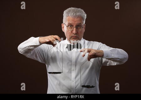 Homme aux cheveux gris âgés de 50 ans, en chemise blanche, lunettes et chapeau pesant quelque chose sur les balances avec petit kettlebells Banque D'Images