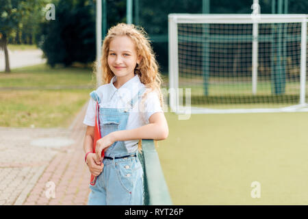 Belle fille blonde aux yeux bleus rire dans un ensemble en denim. pretty student est la tenue d'un dossier rouge avec documents. sweet adolescent se trouve près de Banque D'Images