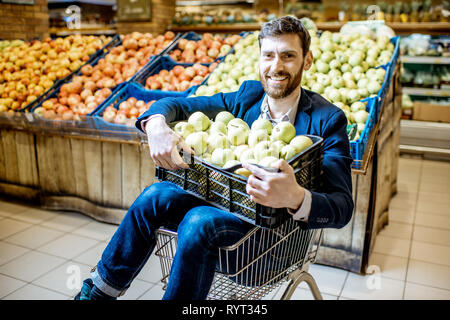 Portrait d'un homme heureux acheteur avec boîte pleine de pommes assis dans le panier dans le supermarché Banque D'Images