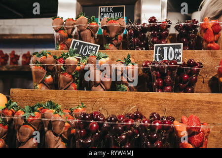 Tasses de fraises, cerises et fraises trempées dans le chocolat au lait en vente, en portions individuelles dans des gobelets en plastique, en haut d'une des étagères en bois. Banque D'Images