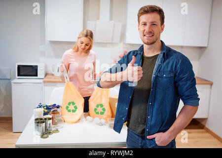 Protéger l'environnement. Young smiling man showing thumb up après le recyclable, tandis que sa femme tenant de sacs à déchets à recycler le symbole à la cuisine Banque D'Images