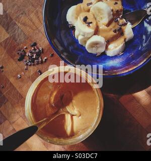 Tranches de banane avec du beurre d'arachide et de cacao nibs en poterie émaillée bleu bol sur billot dans soleil pommelé Banque D'Images