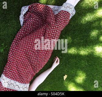 Une jeune femme en robe à motifs rouge se trouve à rêver sous l'ombre des arbres, à côté d'une seule feuille, tombé sur une couverture verte de l'herbe sur une belle journée ensoleillée. Banque D'Images