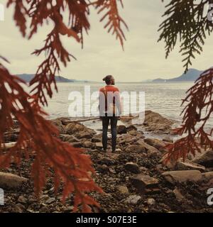 Man on Rocky beach avec chandail orange holding stick orange et branches de cèdre à l'avant-plan. Banque D'Images