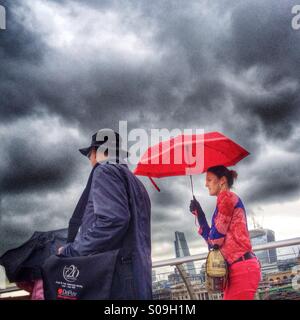 Un homme et une femme colorée en rouge et portant un parapluie rouge se détachent sur un ciel gris foncé Londres pluvieux. Banque D'Images